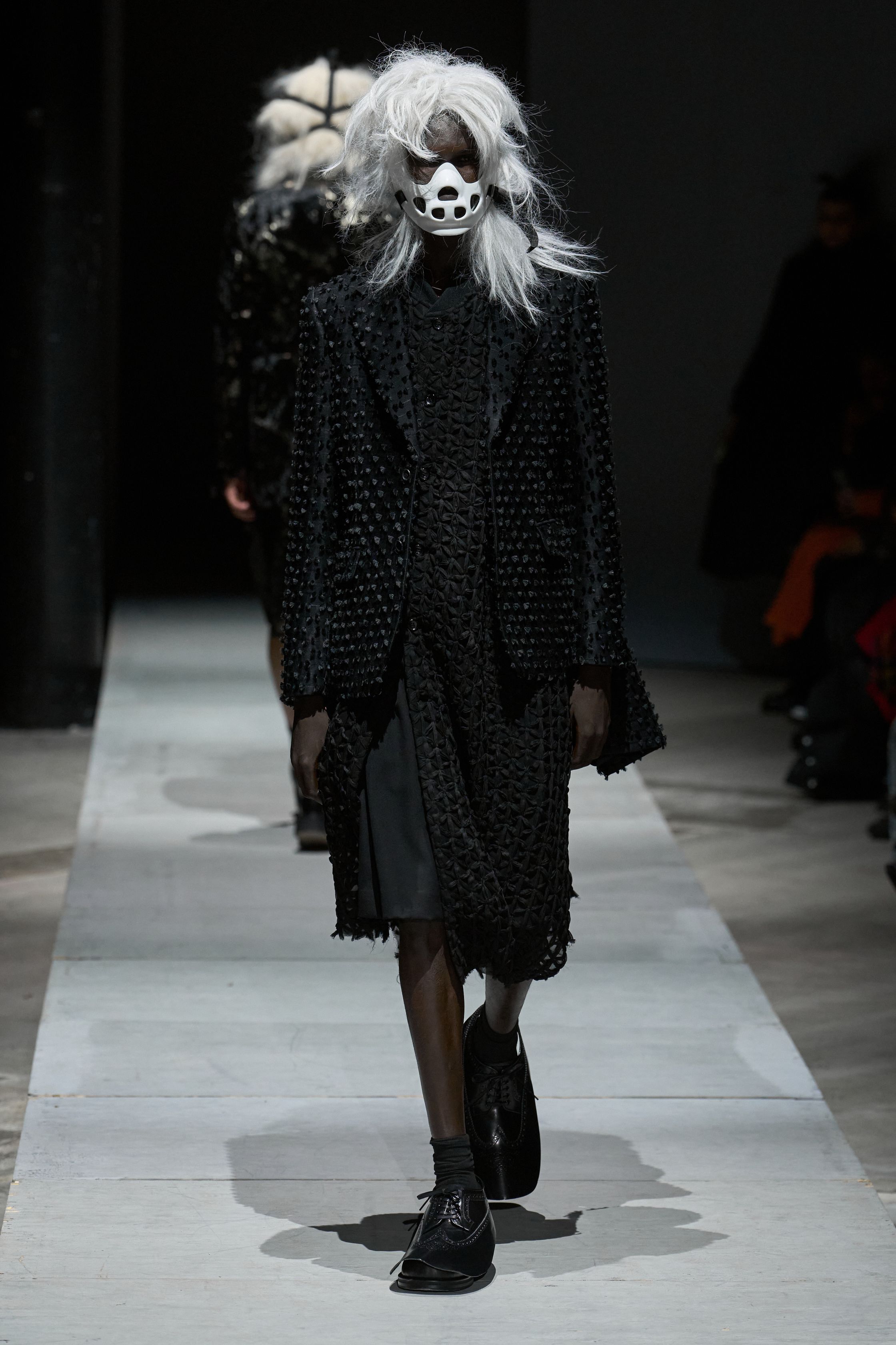 Comme des Garçons Homme Plus 2026秋冬以极端造型继续挑战“男性身体”的社会想象。面具、假发、解构西装与异化比例营造出近乎仪式化的压迫氛围，黑白灰成为思想载体而非装饰。这是一场关于权力、恐惧与集体意识的时装实验，服装不再服务于穿着，而是成为观念的武器-顽石频道趋势研究